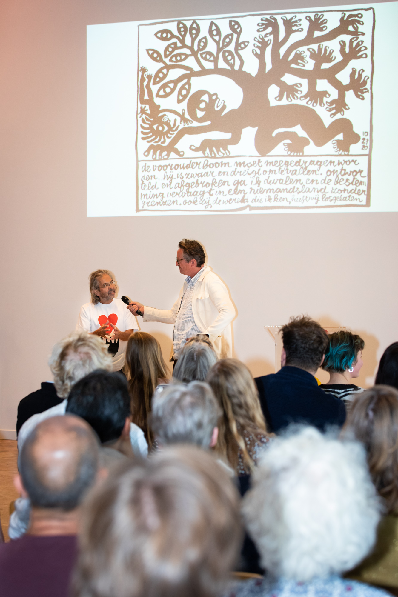 overzichtsfoto van Max Kisman die wordt geïnterviewd door Bart Rutten met op de voorgrond publiek en op de achtergrond een afbeelding van zijn plan
